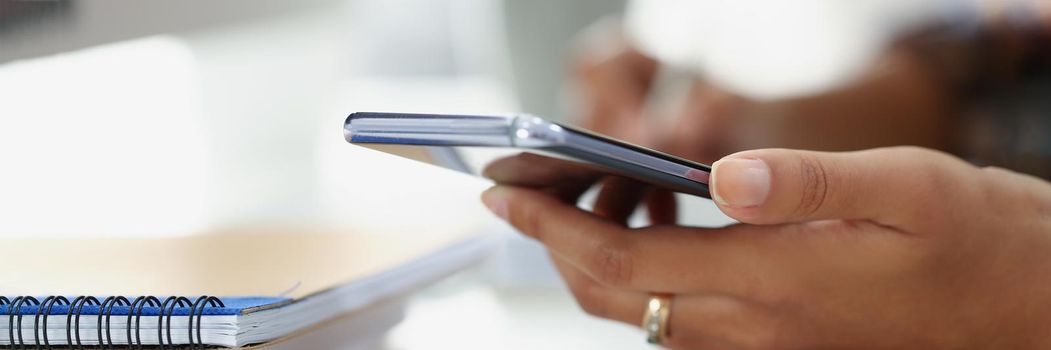 Close-up of woman using modern smartphone while sitting at table with spiral notebook in office. Write in diary and making call. Business, planning concept