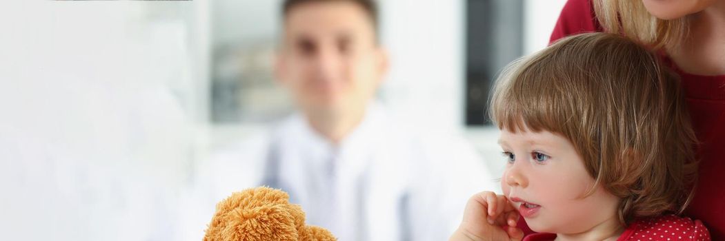 Portrait of little girl with mother play with teddy bear before doctor appointment. Cute child with parent visit family doctor. Hospital, health concept