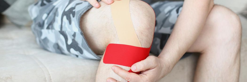 Close-up of person sit on couch with kinesiology tape on knee, pain in leg. Rehabilitation after injury or accident. Physical therapy, healthcare concept