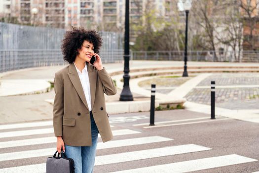 smiling latin business woman walking by the city talking by mobile phone, concept of communication and urban lifestyle, copy space for text