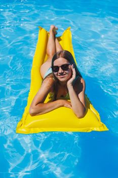 Pretty young woman in swimsuit and sunglasses lying on yellow inflatable mattresses in the pool. Enjoy sunbathing and relaxation.