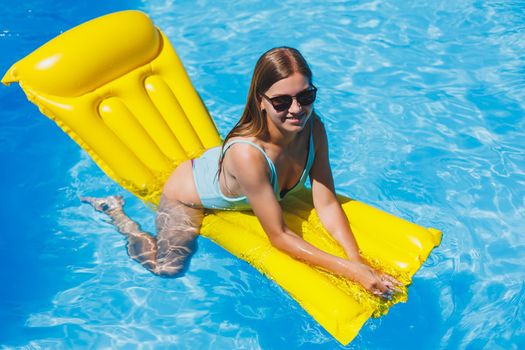 Beautiful young woman in sunglasses on a yellow inflatable mattress in a swimsuit swims in the pool of a luxury hotel, summer vacation, travel, smile