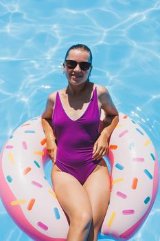 Beautiful slim young woman in a swimsuit enjoying the water park floating in an inflatable big ring on a sparkling blue pool smiling at the camera. Summer vacation.