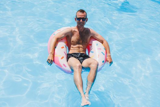 A young man in sunglasses and shorts is relaxing on an inflatable donut in the pool. Summer vacation