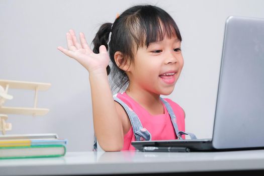 Smiling little Asian girl studying online having video call distant class with teacher using laptop. Happy little girl wave greeting with tutor while studying online with laptop at home. homeschooling