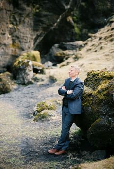 Man in a suit with his arms crossed on his chest stands leaning on a huge boulder. High quality photo