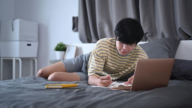 Young man lying on bed and working or studying online with computer laptop.