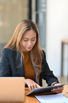 Attractive businesswoman sitting in modern workplace and using digital tablet.