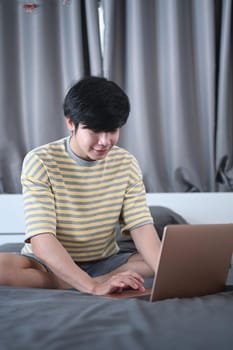 Smiling asian man browsing internet with computer laptop in bedroom.