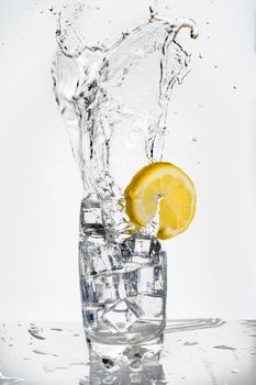 Loud and large splash of an ice cube in a glass of fizzy drink decorated with a lemon. Splash on white background with reflection on the base. vertical format. Hard ligth