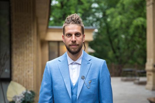 Portrait of a young man 25 years old with a hipster hairstyle and beard in a blue three-piece suit in a restaurant space in a park area.