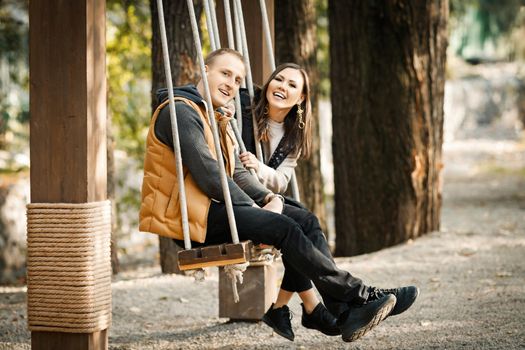 Side view of young cute couple riding on swing in autumn in warm clothes of basic colors in park outdoors. High quality photo
