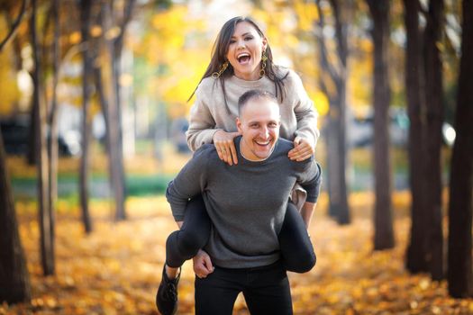 A happy man carries a laughing woman on his back to enjoy the last warm sunny autumn days together.