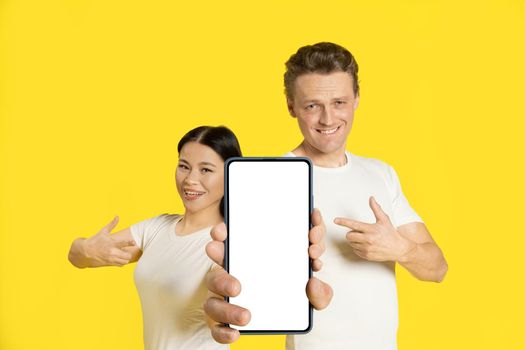 Holding together smartphone with white screen caucasian man and asian woman pointing finger excited new mobile app advertisement isolated on yellow background. Product placement.
