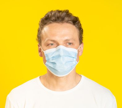 Sad face young man wearing a medical mask on his face. Sick young man in face mask wearing white t-shirt isolated on yellow background. A sick caucasian man in protective face mask.