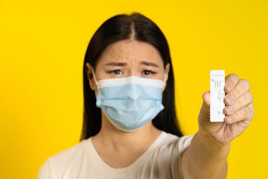 Asian woman in medical face mask and test in hand sad looking at camera isolated on yellow background. Pandemic concept, monkey pox or coronavirus, healthy lifestyle. Selective focus on test.