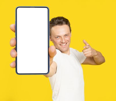 Holding huge smartphone with white screen handsome blond man pointing at white empty screen, wearing white t-shirt and jeans isolated on yellow background. Mobile app advertisement, great offer.