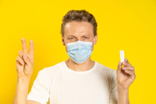 Happy young man gesturing v sign holding test in hand wearing medical face mask celebrating victory pandemic coronavirus or monkeypox. Man in white t-shirt and medical mask on yellow background.