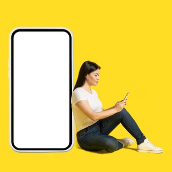 Sitting on the floor middle aged asian woman with phone in hands leaned on giant, huge smartphone with white screen wearing white t-shirt and jeans isolated on yellow background. Free space mock up.