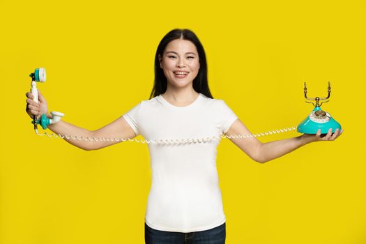 Happy asian woman sideways stretched cable of old fashion telephone looking at camera isolated on yellow background. Beautiful woman in casual look. Telecommunications concept.