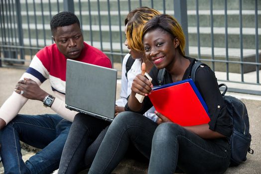 Group of attractive smiling young casual dressed students using laptop sitting outdoors on campus of university.