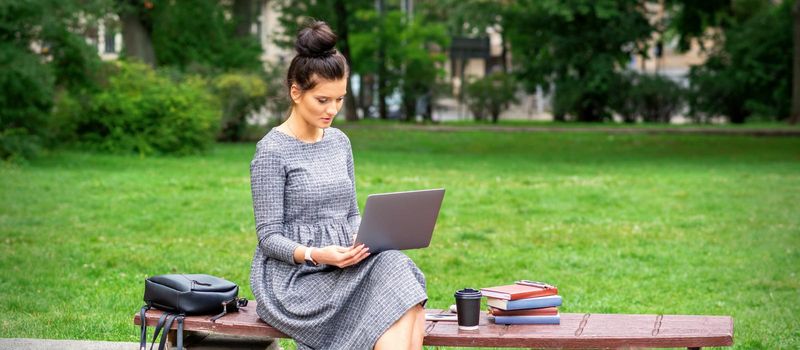 Beautiful young woman sitting on a bench with a laptop and working or studying outdoors