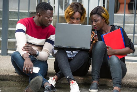 Group of attractive smiling young casual dressed students using laptop sitting outdoors on campus of university.