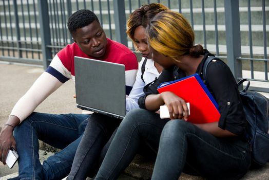 Group of attractive smiling young casual dressed students using laptop sitting outdoors on campus of university.