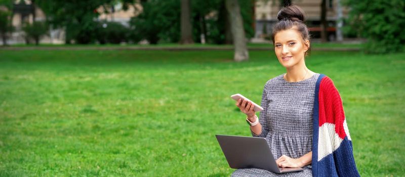 Beautiful smiling young woman sitting on the bench with smartphone and laptop in the park