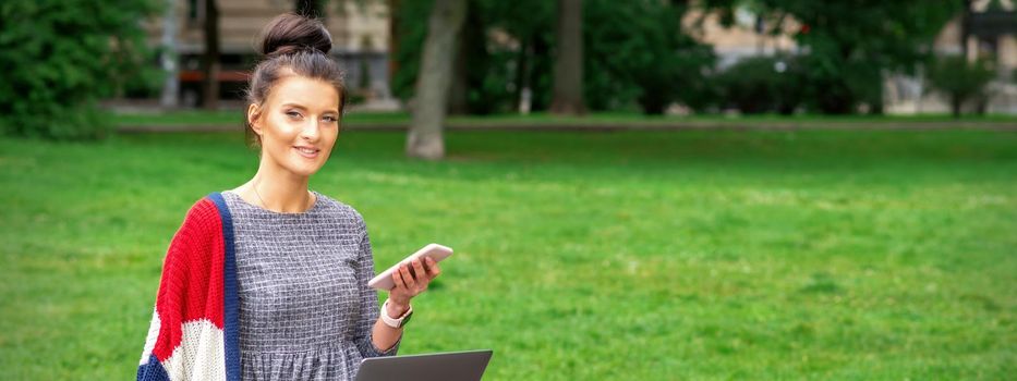 Beautiful smiling young woman sitting on the bench with smartphone and laptop in the park