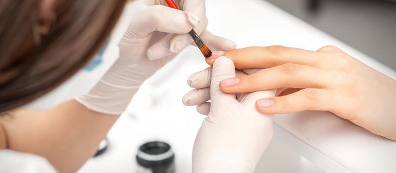 Manicure master is covering female nails with transparent nail polish in a nail salon