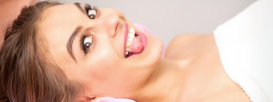 Beautiful young caucasian woman sticking out tongue and lying in spa beauty salon