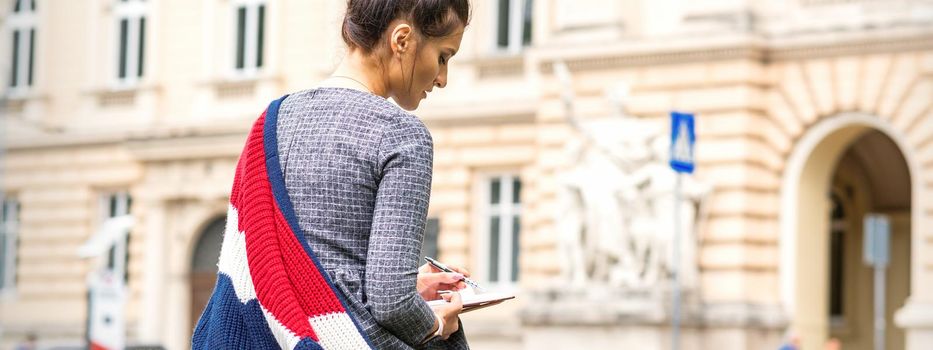 The female student writes into a notebook sitting on the bench against the background of the university