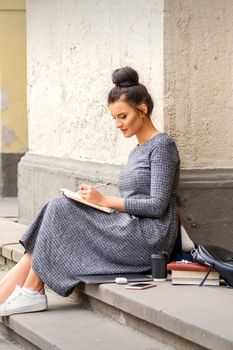 Female student in long dress reading a book on stairs of university building outdoors