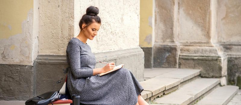 Female student in long dress reading a book on stairs of university building outdoors