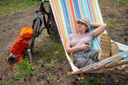 Caucasian woman drinks hot tea from a thermo mug while lying in a hammock in nature