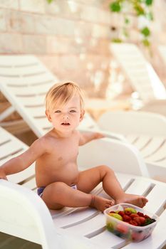 Little girl sits on a white sun lounger in front of a fruit box. High quality photo