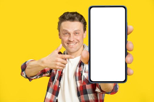 Pointing at giant, huge smartphone with white empty screen blond man, wearing red plaid shirt. Man with phone display mock up isolated on yellow background. Mobile app advertisement.