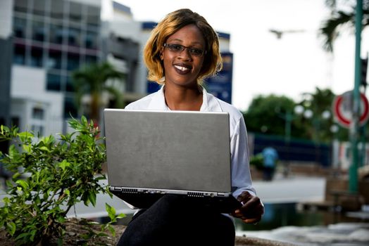 young happy female student wearing glasses sitting outside and in the city and working on laptop