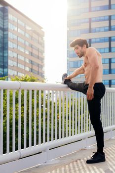vertical photo of a handsome shirtless sportsman stretching his leg muscles in front of city buildings, concept of urban sport and healthy lifestyle, copy space for text