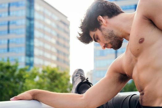 handsome shirtless sportsman stretching his leg muscles in front of city buildings, concept of urban sport and healthy lifestyle, copy space for text