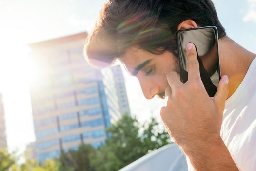 handsome young man talking on his mobile phone in front of city buildings at sunset, technology concept and urban lifestyle, copy space for text