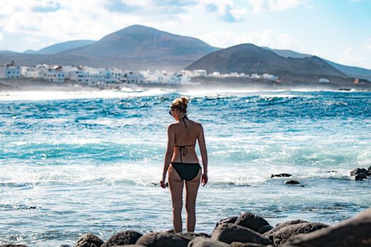 Female tourist at wild rocky beach and coastline of surf spot La Santa Lanzarote, Canary Islands, Spain. La Santa village and volcano mountain in background