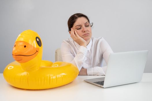 Caucasian woman sits at a table with a laptop and an inflatable duck on a white background. Office worker dreaming of vacation