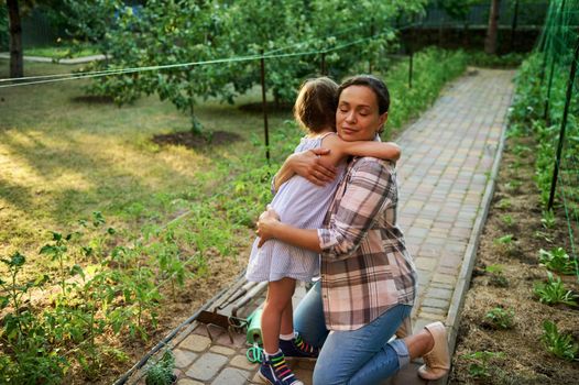 Multiethnic charming woman, loving mother hugging her lovely little daughter, standing together in the eco farm, planting and caring for seedlings. Install love and care for nature since childhood