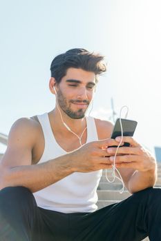 vertical photo of a handsome sportsman with earphones listening to music and consulting his phone, concept of technology and healthy lifestyle, copy space for text