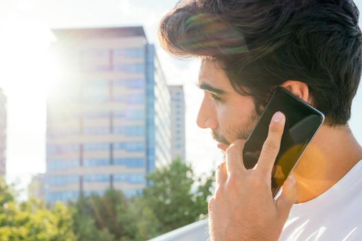 handsome young man talking on his smartphone in front of city buildings at sunset, technology concept and urban lifestyle, copy space for text