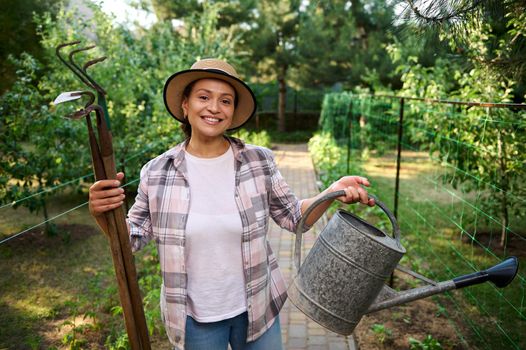 Charming Caucasian woman farmer with garden tools, smiles toothy smile looking at camera, while working in organic farm, inspecting planted seedlings and cultivating ecologically friendly vegetables