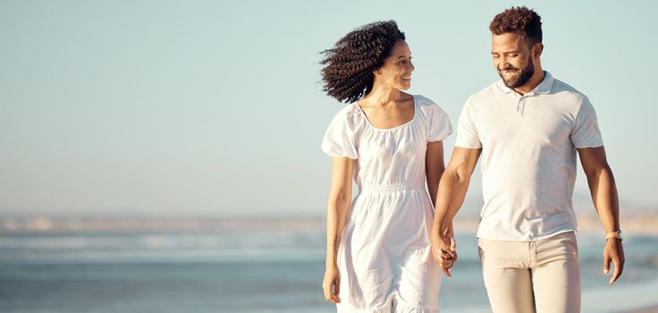Happy mixed race young couple holding hands while walking on the beach together. Hispanic couple traveling and enjoying vacation and being romantic on the beach.