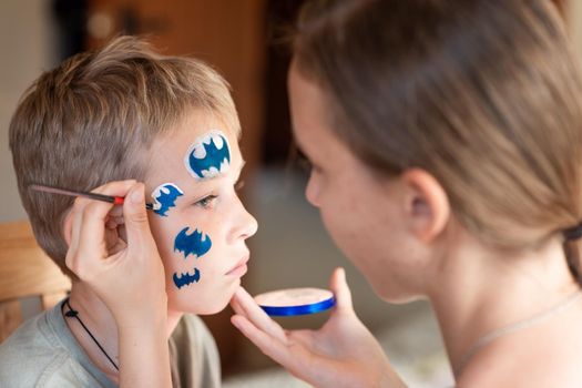 Child animator, artist's hand draws face painting to little boy. Child with funny face painting. Painter makes blue batman mask on boy's face. Face painting at children holiday, event, birthday party, entertainment.